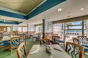 En restaurang med bord och stolar, stora fönster med utsikt över havet och ett tak med infälld belysning.