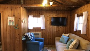L’intérieur d’un chalet accueillant, avec des murs en bois, un fauteuil bleu, un canapé beige, un téléviseur à écran plat et une fenêtre avec des rideaux.
