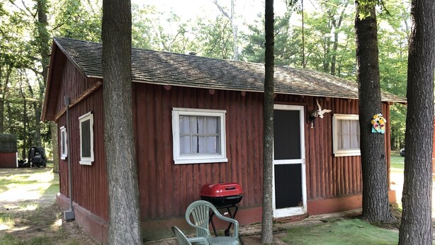 Une petite cabane en bois avec une seule porte et deux fenêtres, un barbecue rouge à l’extérieur et une chaise en plastique devant.