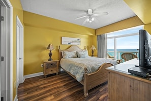 A bedroom with a wooden bed, a nightstand with lamps, a TV, and a view of the ocean.