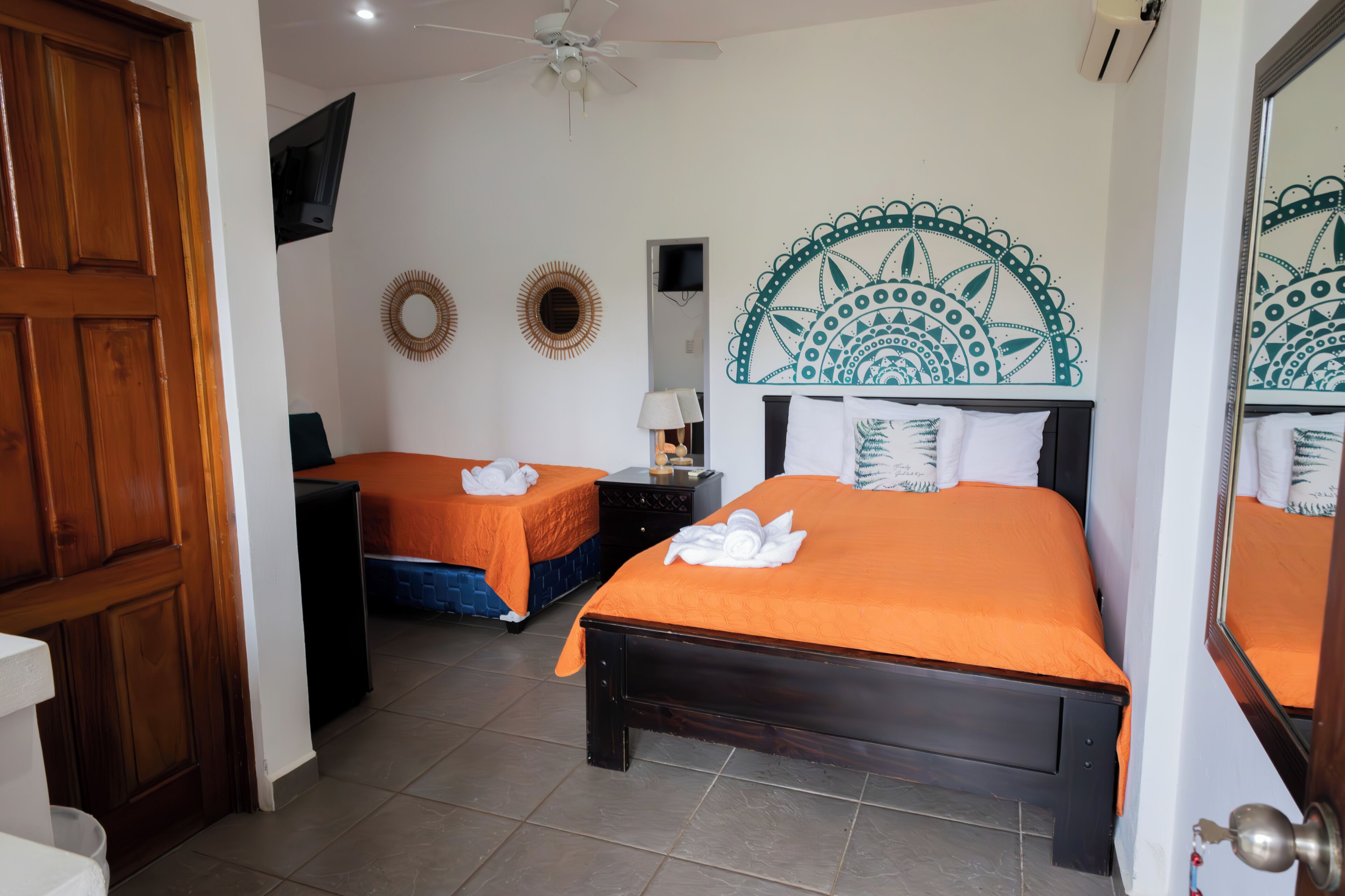 A hotel room with two beds, a wooden door, a ceiling fan, and a wall-mounted TV.
