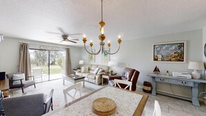 A spacious living room with a ceiling fan, a chandelier, a dining table with a woven placemat, a sofa, a leather armchair, and a painting on the wall. - Ponce Landing 5 - Ocean front 2 bedroom townhome with garage (St. Augustine)