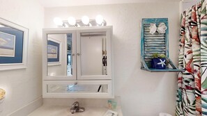 A bathroom with a vanity, a mirror with lights, a shelf with decorative items, and a curtain with a tropical pattern. - Ponce Landing 5 - Ocean front 2 bedroom townhome with garage (St. Augustine)