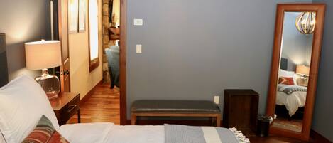 Un dormitorio con cama, mesita de noche, lámpara, banqueta y espejo.