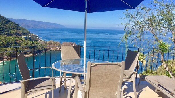 Un balcone con tavolo e sedie in vetro, un ombrellone blu e vista sull'oceano e sulle colline.