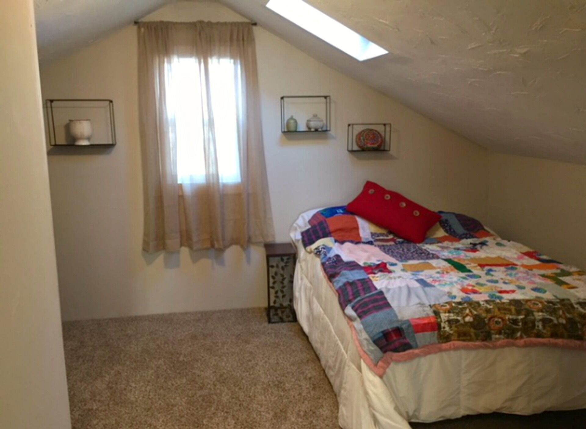 Une chambre avec un plafond en pente, un lit recouvert d’une couette aux couleurs vives, un oreiller rouge et une fenêtre ornée de rideaux légers.