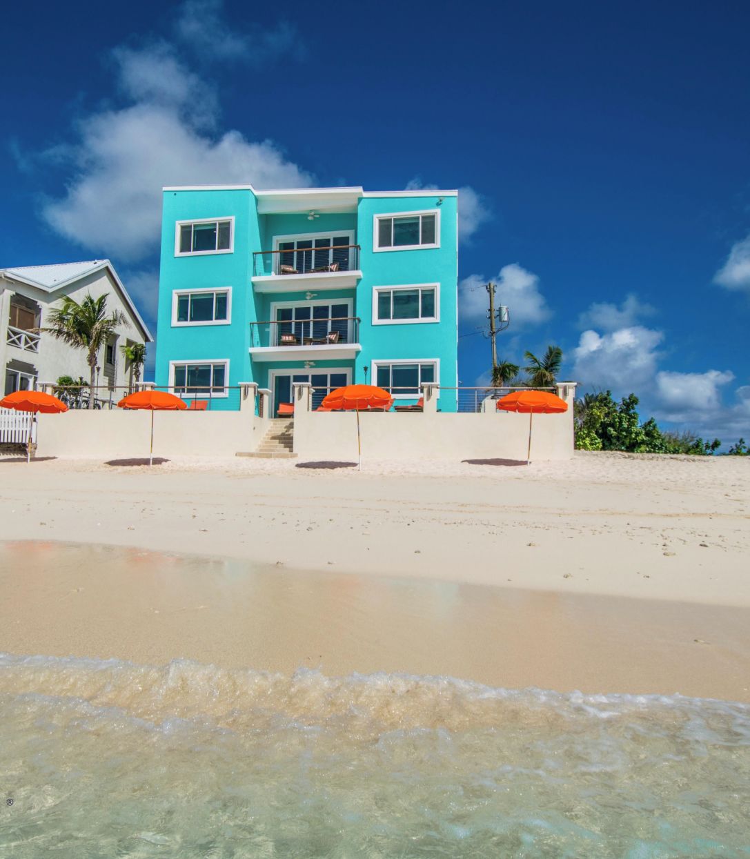 A two-story building with a light blue facade, white trim, and multiple balconies. There are orange umbrellas and a white fence in the foreground.