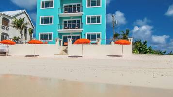 A two-story building with a light blue facade, white trim, and multiple balconies. There are orange umbrellas and a white fence in the foreground.