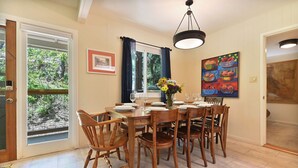 Un'area pranzo con un tavolo di legno apparecchiato per sei persone, circondato da sei sedie di legno. Si nota una finestra con tende, un quadro appeso alla parete e una porta che dà su un'altra stanza.