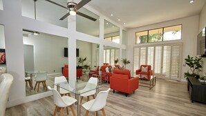 Un vaste espace de vie comprenant une table à manger en verre, des chaises blanches, des canapés rouges et un ventilateur de plafond.