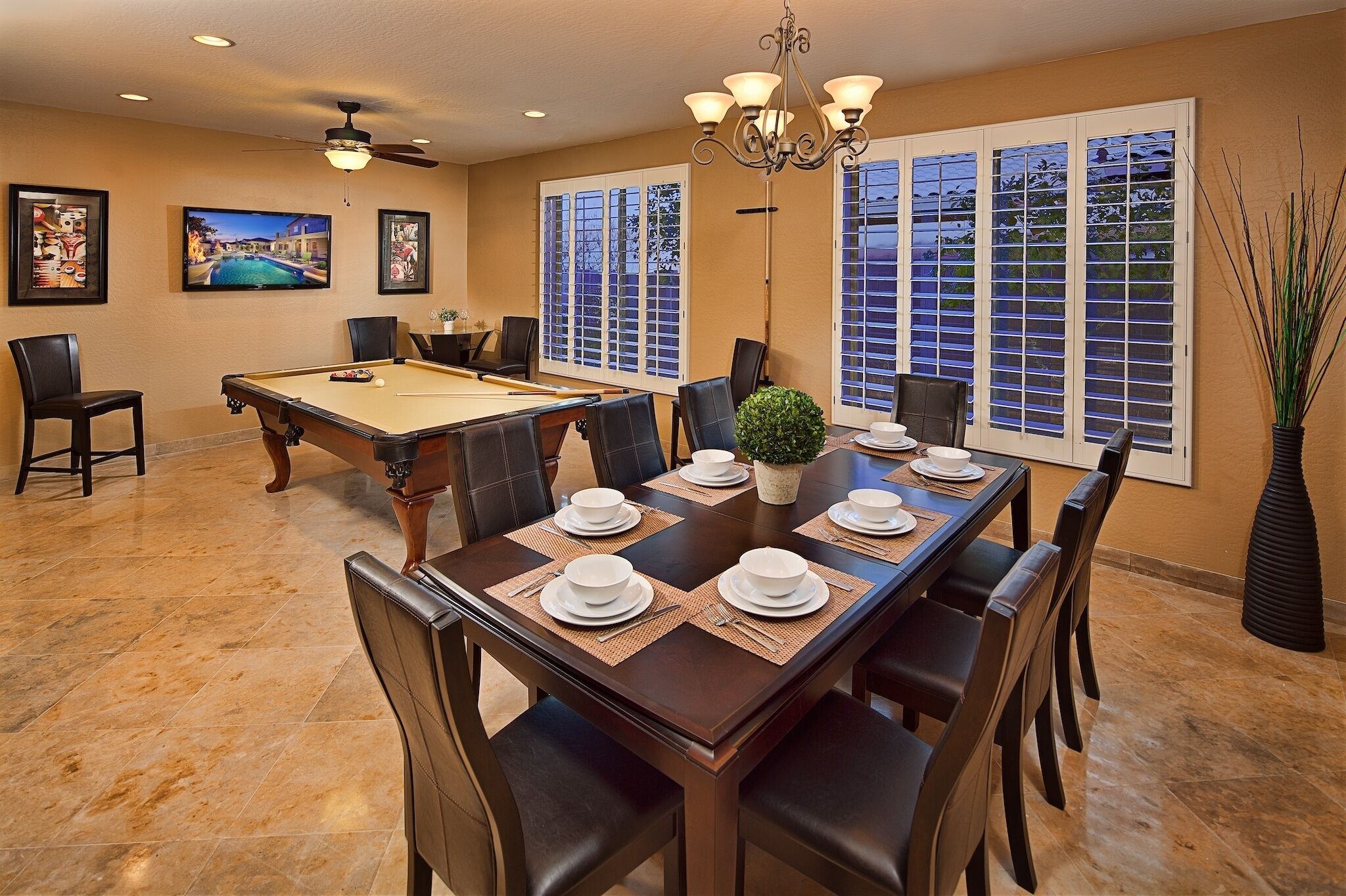 A dining room with a pool table, a chandelier, and large windows with blinds.