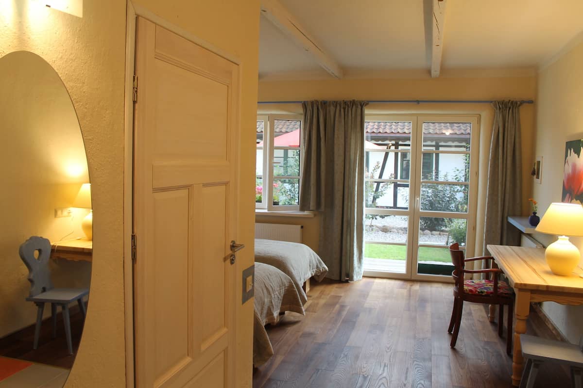 A room with a bed, a table with a lamp, a chair, and a door leading to an outdoor area with a view.
