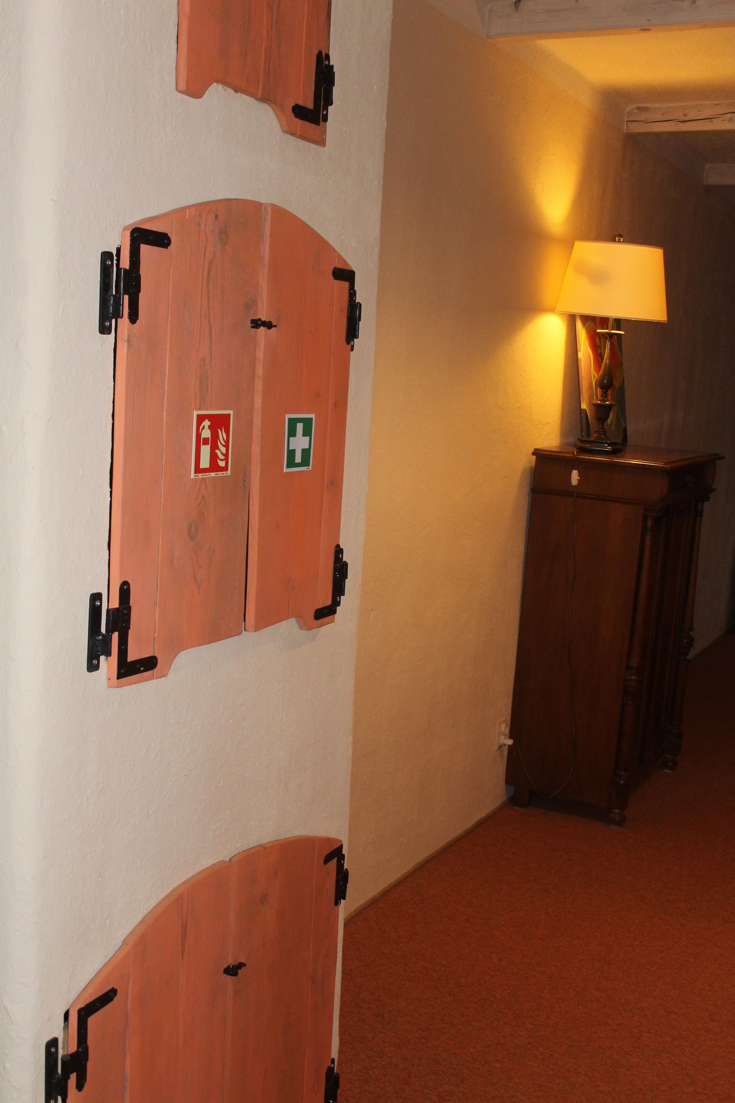 En hall med dörrar av trä, försedda med symboler för nödsituationer, en lampa och ett träskåp.