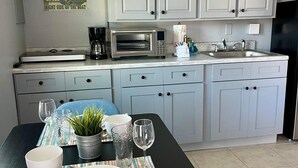 Een moderne keuken met grijze kastjes, een magnetron en een spoelbak. Er staat een eettafel met een gestreepte placemat, een kom, een glas en een plantje.