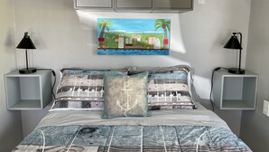 Een slaapkamer met een bed, twee nachtkastjes met lampen, een wandplank en een schilderij van een strandgezicht.
