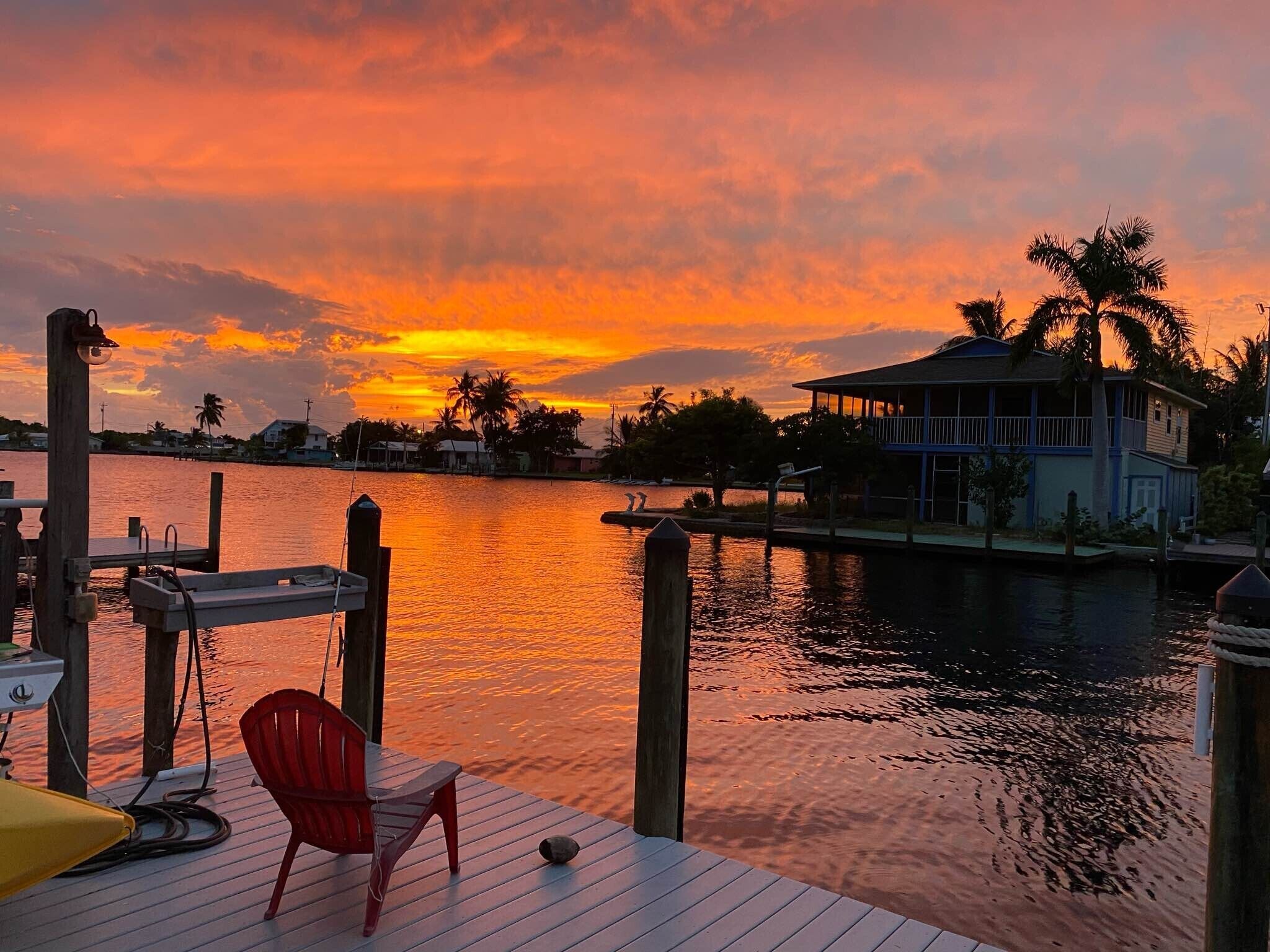 A wooden dock with a red chair and a yellow umbrella, overlooking a calm body of water with a house and palm trees in the background.