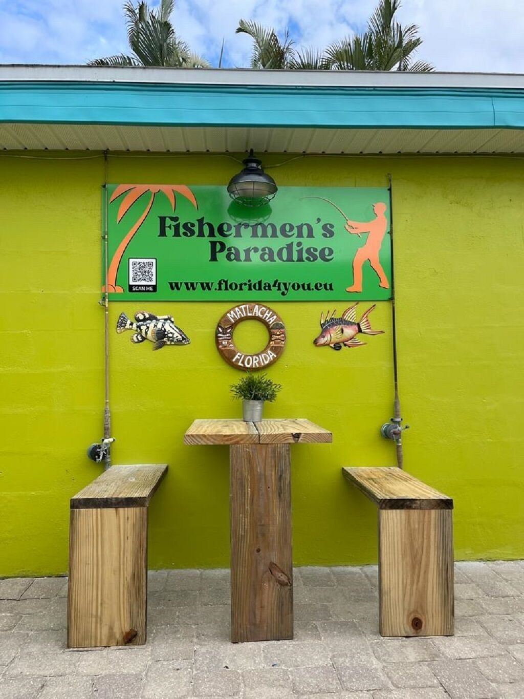 A wooden table with two stools in front of a green wall with a sign reading "Fishermen's Paradise" and a website URL.