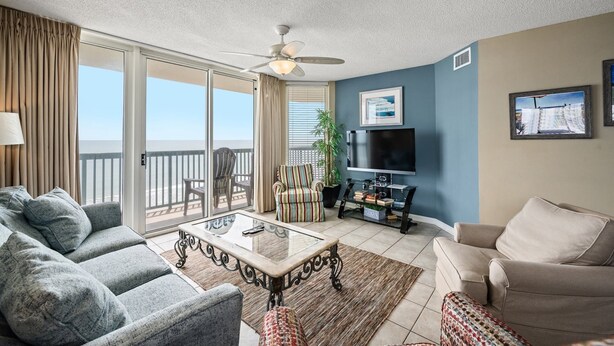 Un salon avec balcon, un ventilateur de plafond, un téléviseur à écran plat et une grande fenêtre avec des rideaux.