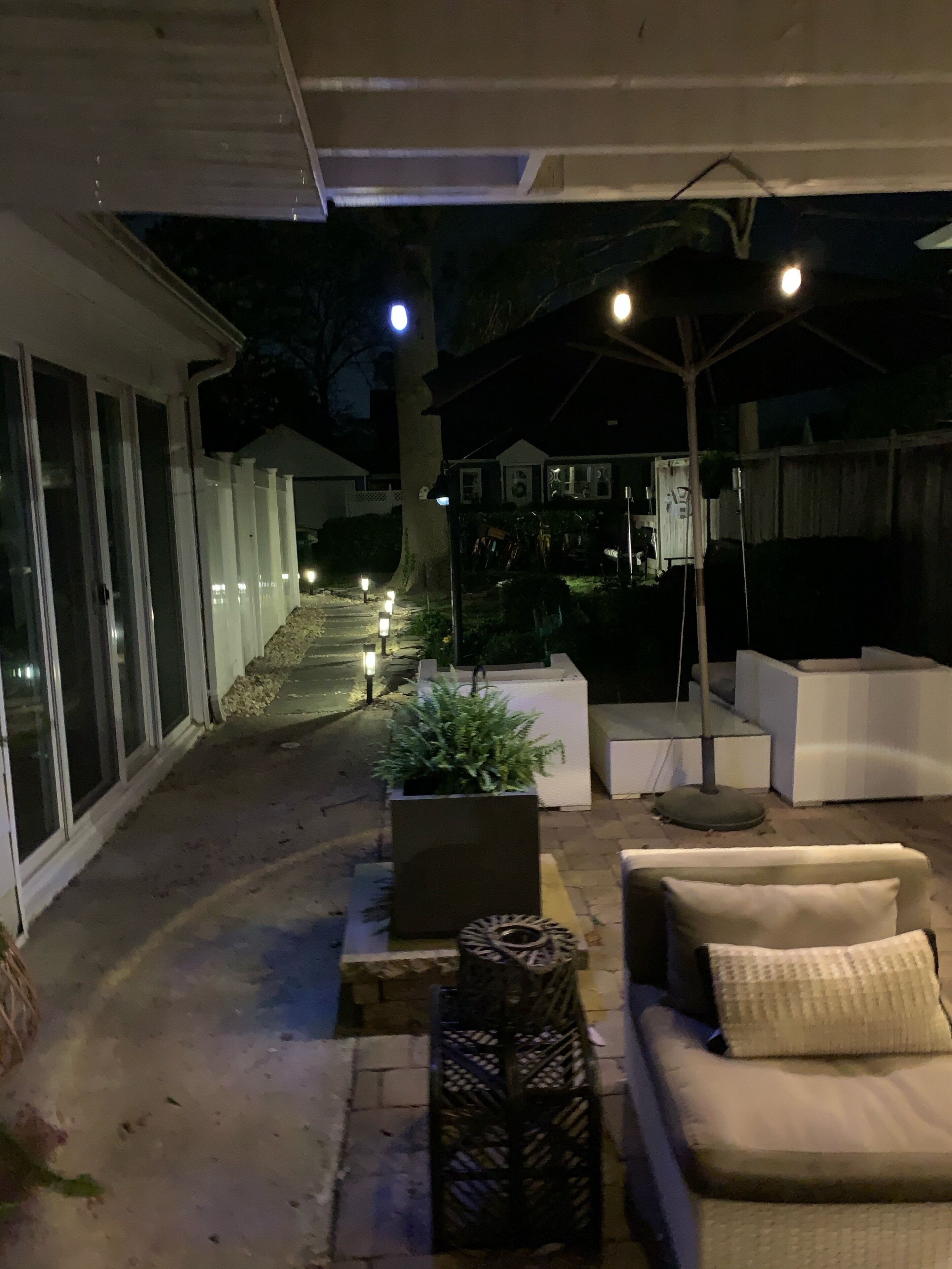 An outdoor patio area with a white sofa, a small table, and potted plants. There is a pathway leading to a house with a covered porch.