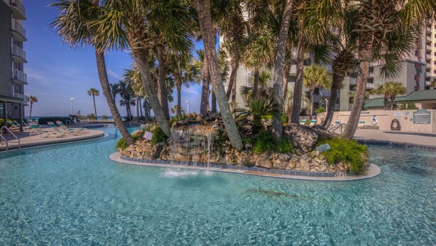 A clear pool with a waterfall feature surrounded by palm trees, lounge chairs, and a view of a high-rise building.
