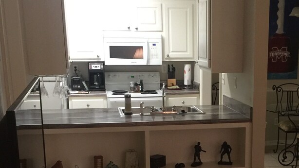 A kitchen with white cabinets, a microwave, and a coffee maker. There is a shelf with decorative items and a small table with a remote control. - Road Dawg Condo  Welcome to God's country.  (Starkville)