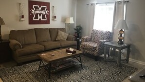 A living room with a brown sofa, a wooden coffee table, patterned rugs, a ceiling fan, and a wall-mounted TV. - Road Dawg Condo  Welcome to God's country.  (Starkville)