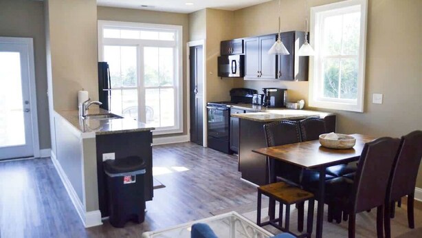 A modern kitchen with dark cabinets, a central island, and a dining area with a wooden table and chairs. - 3BR Waterfront Home Fishing Boating Views (Stacy)