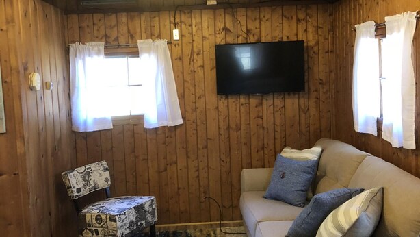 Ett mysigt rum med träväggar, en soffa, en mönstrad fåtölj, en platt-TV och en blå matta.
