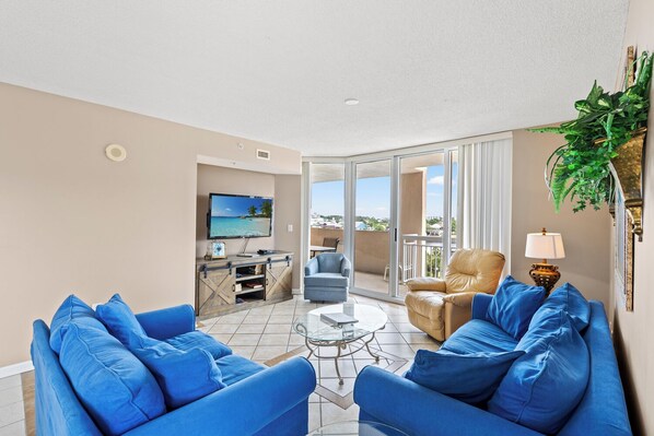 A spacious living room with a TV, blue sofas, a coffee table, and a balcony with a view.