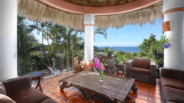 A covered outdoor seating area with wicker furniture, a wooden table, and a view of the ocean. - Caracol Estates (Sayulita)