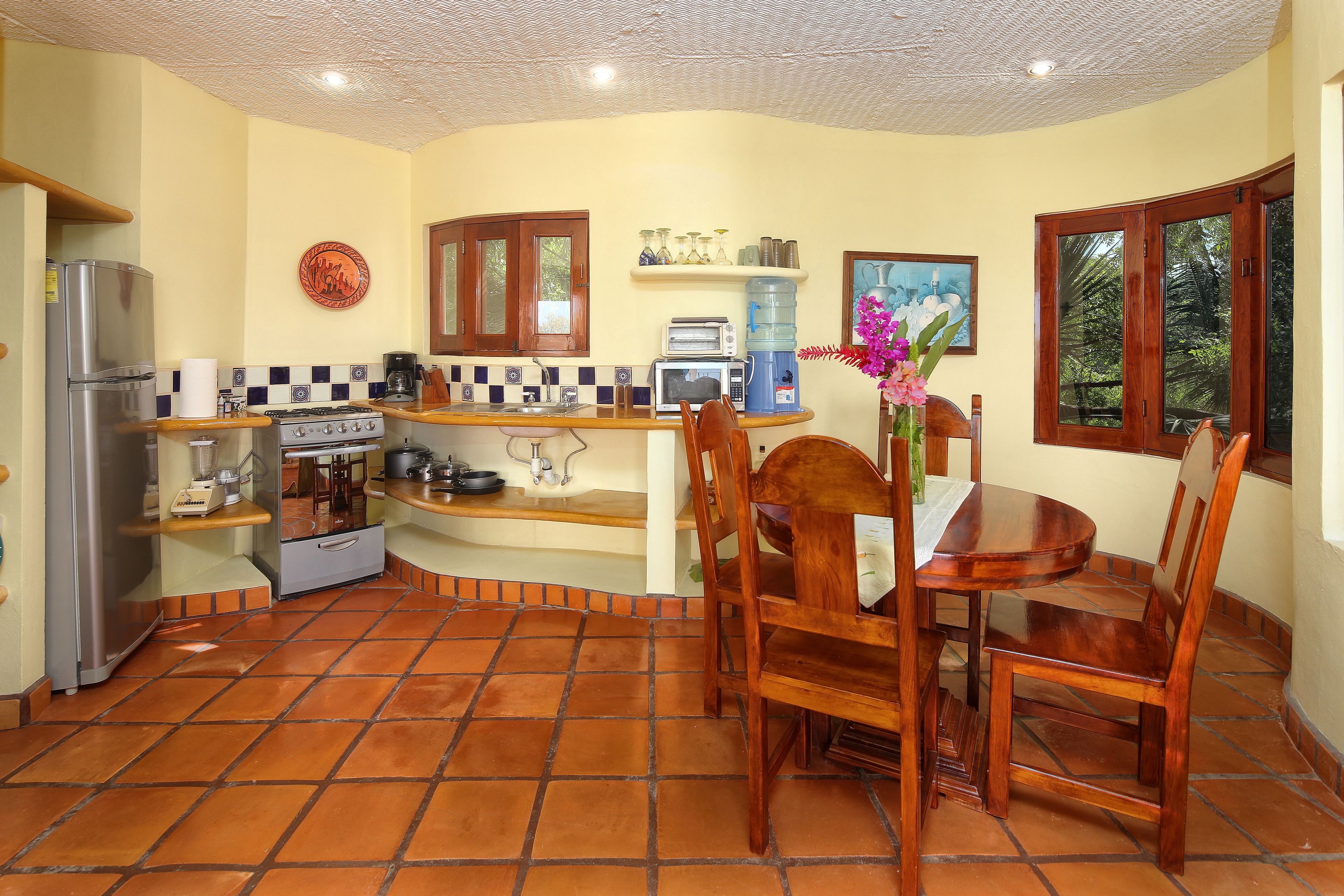A compact kitchen with a refrigerator, stove, microwave, and coffee maker. There is a dining table with chairs and a window with a view of greenery.