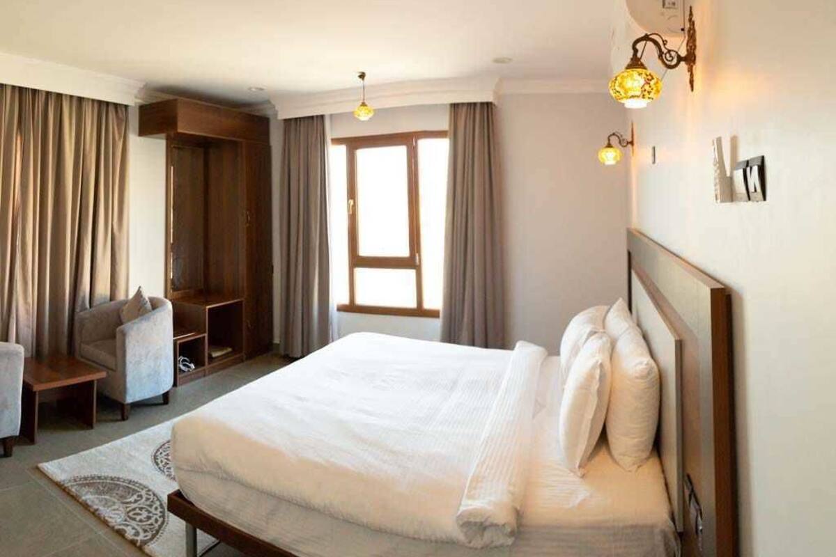A bedroom with a large bed, a wooden wardrobe, a seating area with a chair, and a window with curtains.
