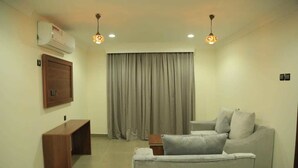 A living room with a sofa, a wooden bench, a TV mounted on the wall, and a large window with curtains. - Nizwa Residence Hotel Apartment (Nizwa)