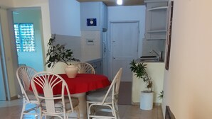 A dining area with a table covered by a red tablecloth, surrounded by white chairs. There are potted plants on the table and a white cabinet in the background.