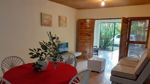 A dining area with a red tablecloth, white chairs, and a potted plant. There is a wooden wall and a sliding door leading to a balcony with a white sofa.