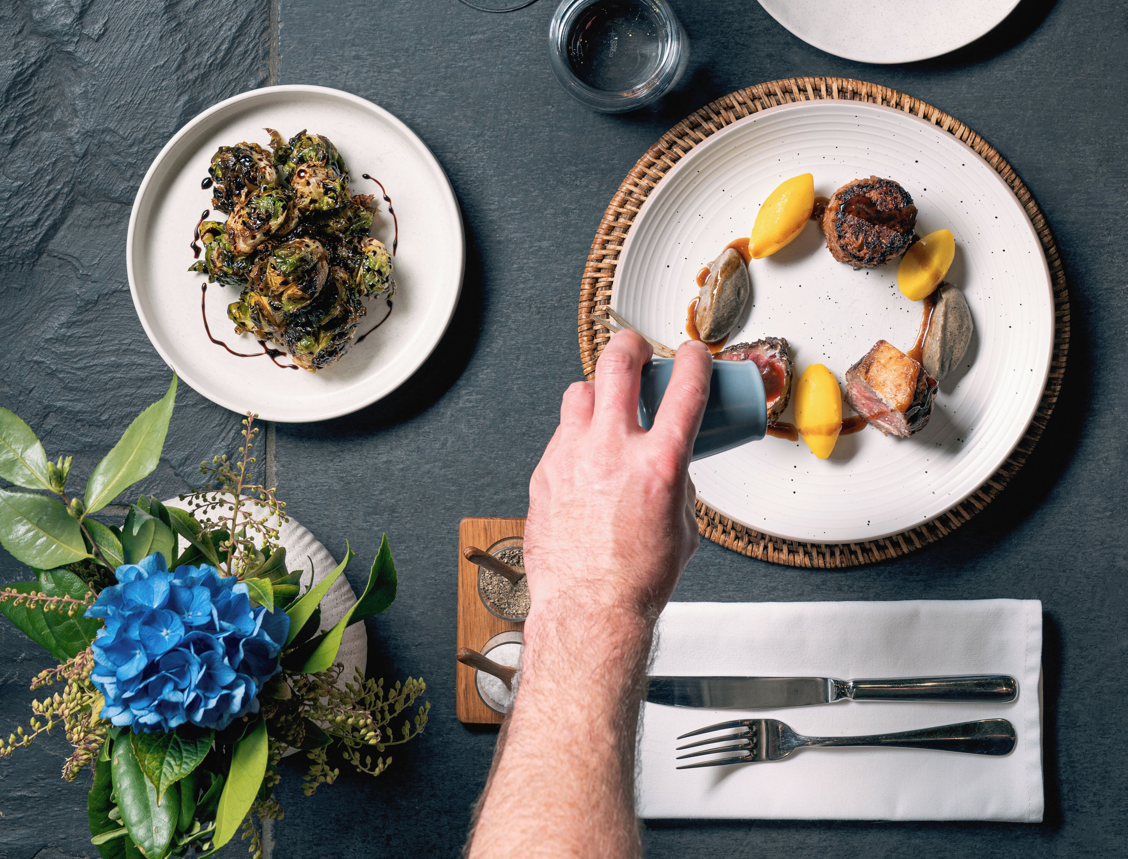 A dining table with plates of food, a hand reaching for a fork, and a napkin with cutlery.