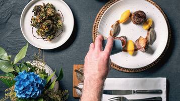 A dining table with plates of food, a hand reaching for a fork, and a napkin with cutlery.