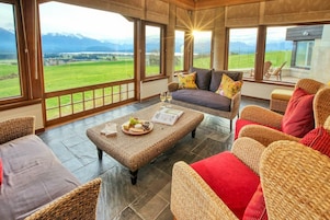 Amplia habitación con muebles de mimbre, una mesa de comedor con una bandeja de quesos y grandes ventanales con vistas a un paisaje verde y montañas.