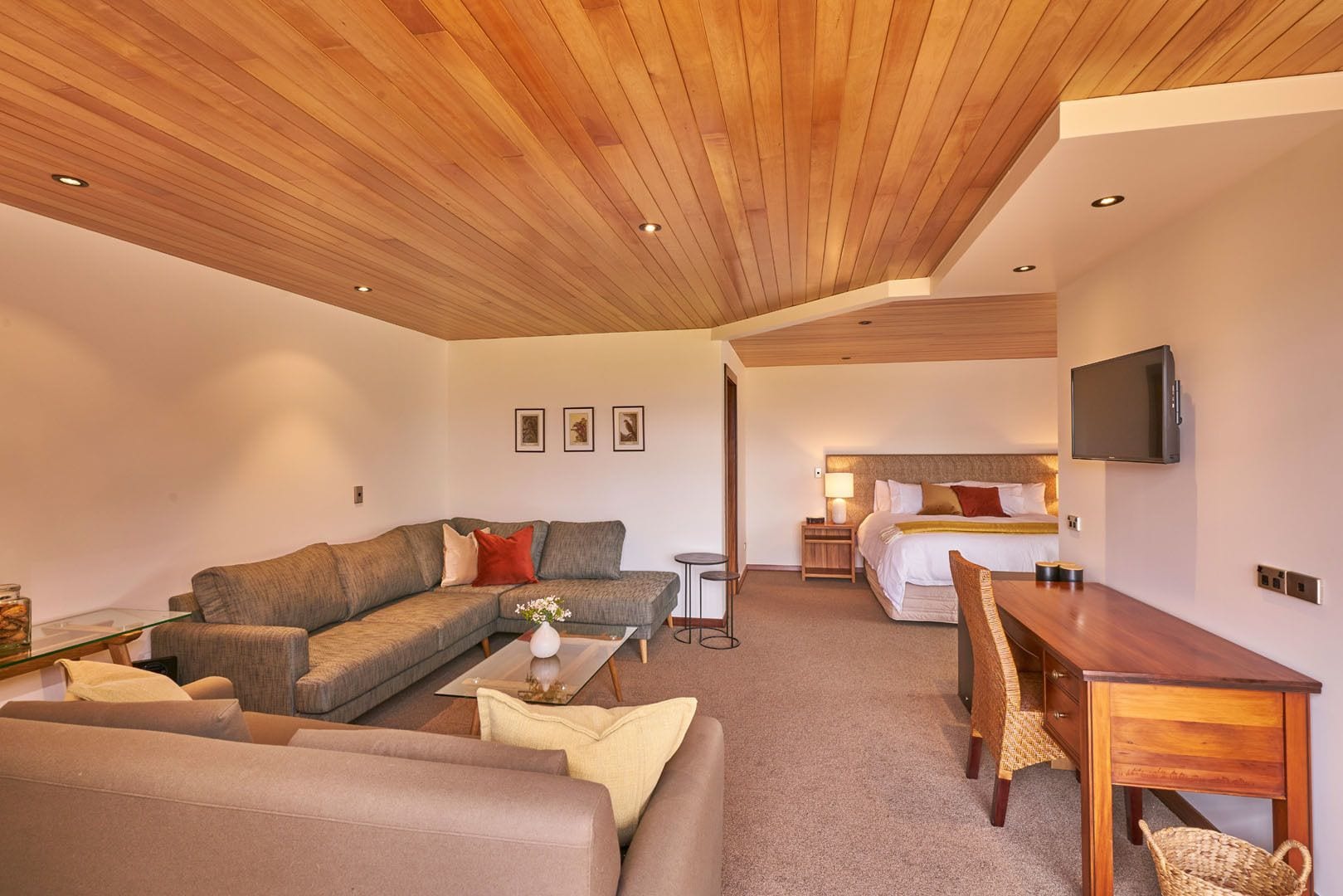 A modern living room with a wooden ceiling, a sofa, a coffee table, a TV, and a bed with a headboard.