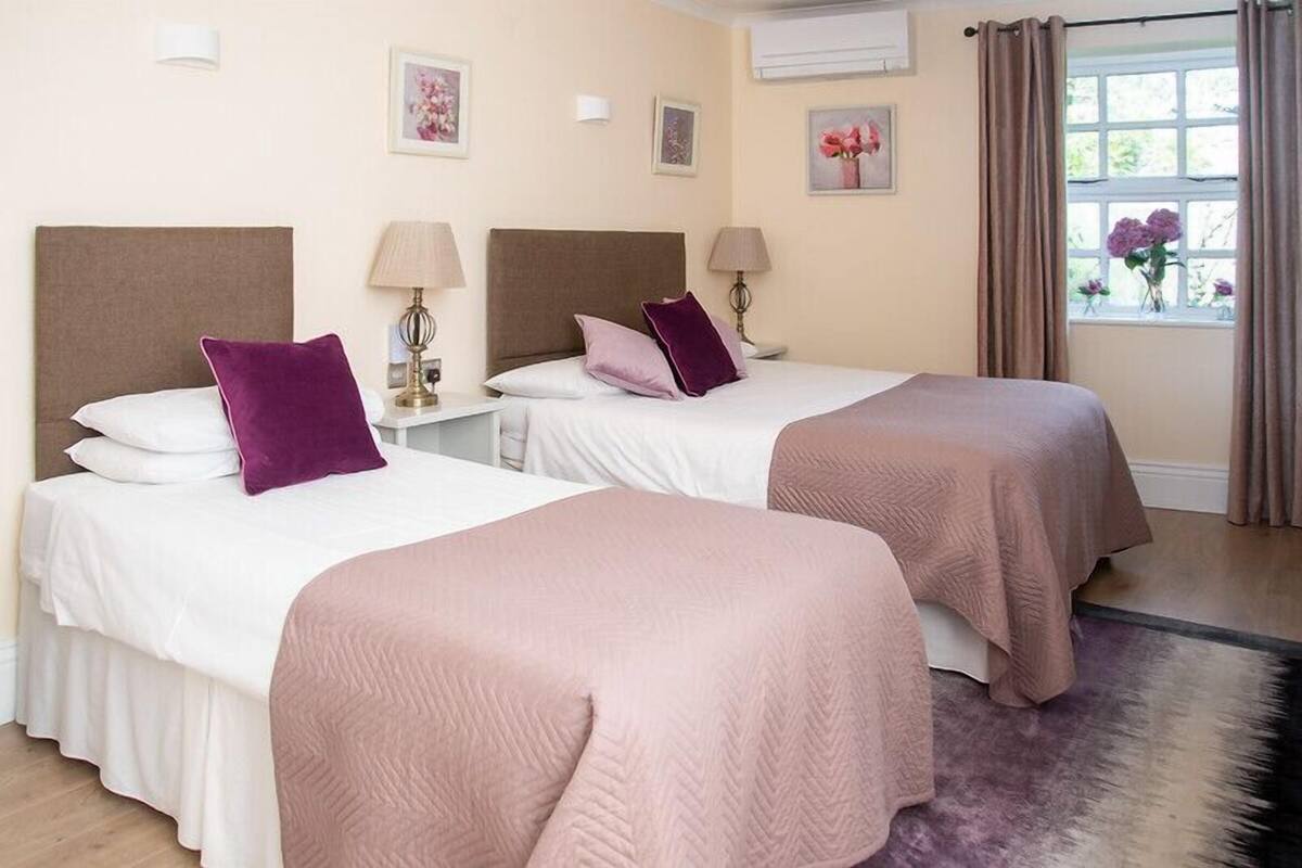A hotel room with two beds, each with a headboard, a nightstand with a lamp, and a window with curtains.