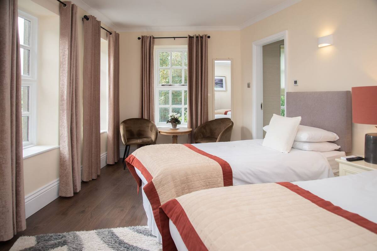 A hotel room with two beds, a small table with a plant, two chairs, and a window with curtains.