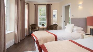 A hotel room with two beds, a small table with a plant, two chairs, and a window with curtains.
