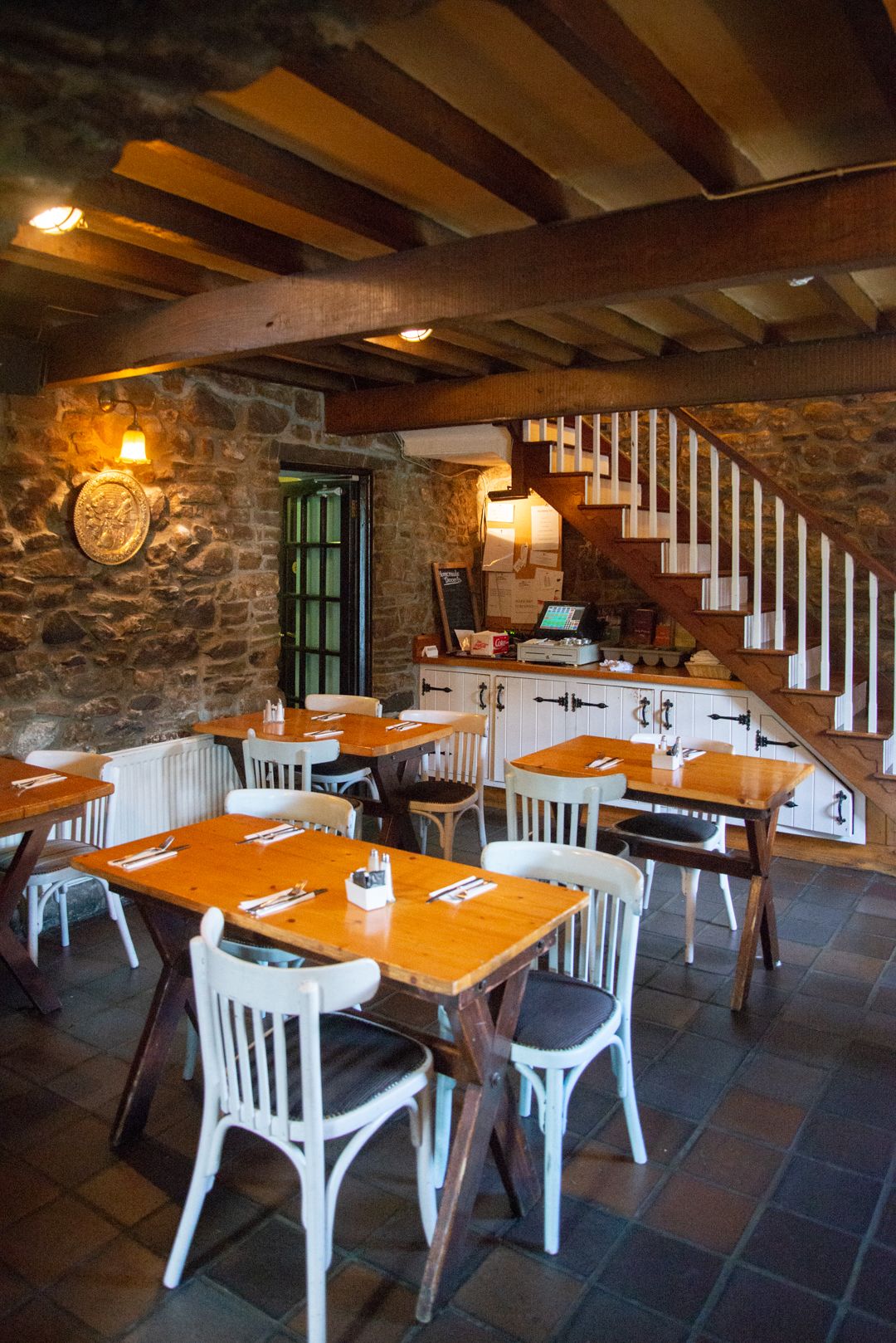 Un restaurante con paredes de piedra, mesas de madera y sillas blancas. Se ve una escalera con barandillas de madera que sube a un nivel superior.