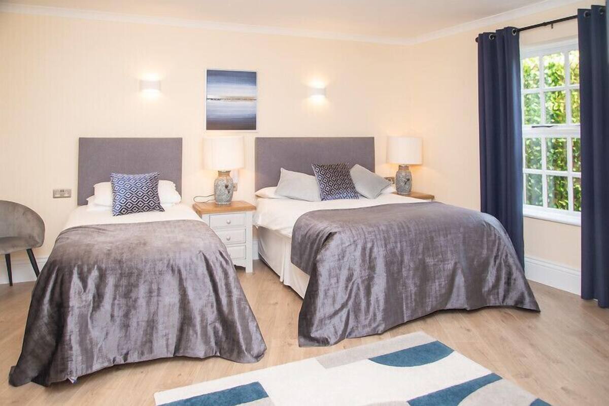 A double room with two beds, each with a grey bedspread, bedside tables with lamps, and a large window with curtains.