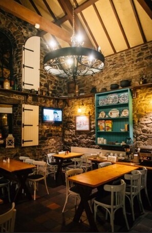 Interior rústico de restaurante con paredes de piedra, mesas de madera y un televisor montado en la pared.