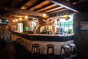 Um bar com parede de pedra, balcão de madeira, banquetas e luminárias pendentes.