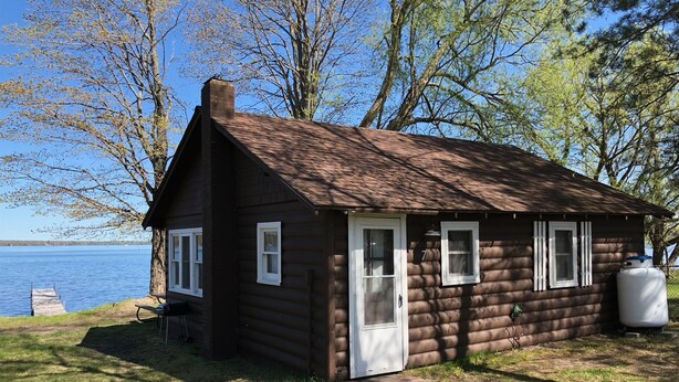 A small log cabin with a white door, two windows, and a chimney. It is situated by a lake with trees in the background.