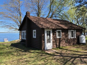 A small log cabin with a white door, two windows, and a chimney. It is situated by a lake with trees in the background. - Cabin #7 - "Waugoshance" (Carp Lake)