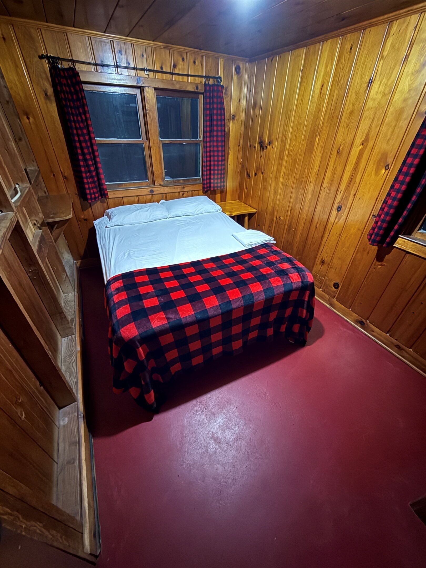 A wooden cabin room with a bed, two windows, and plaid curtains.