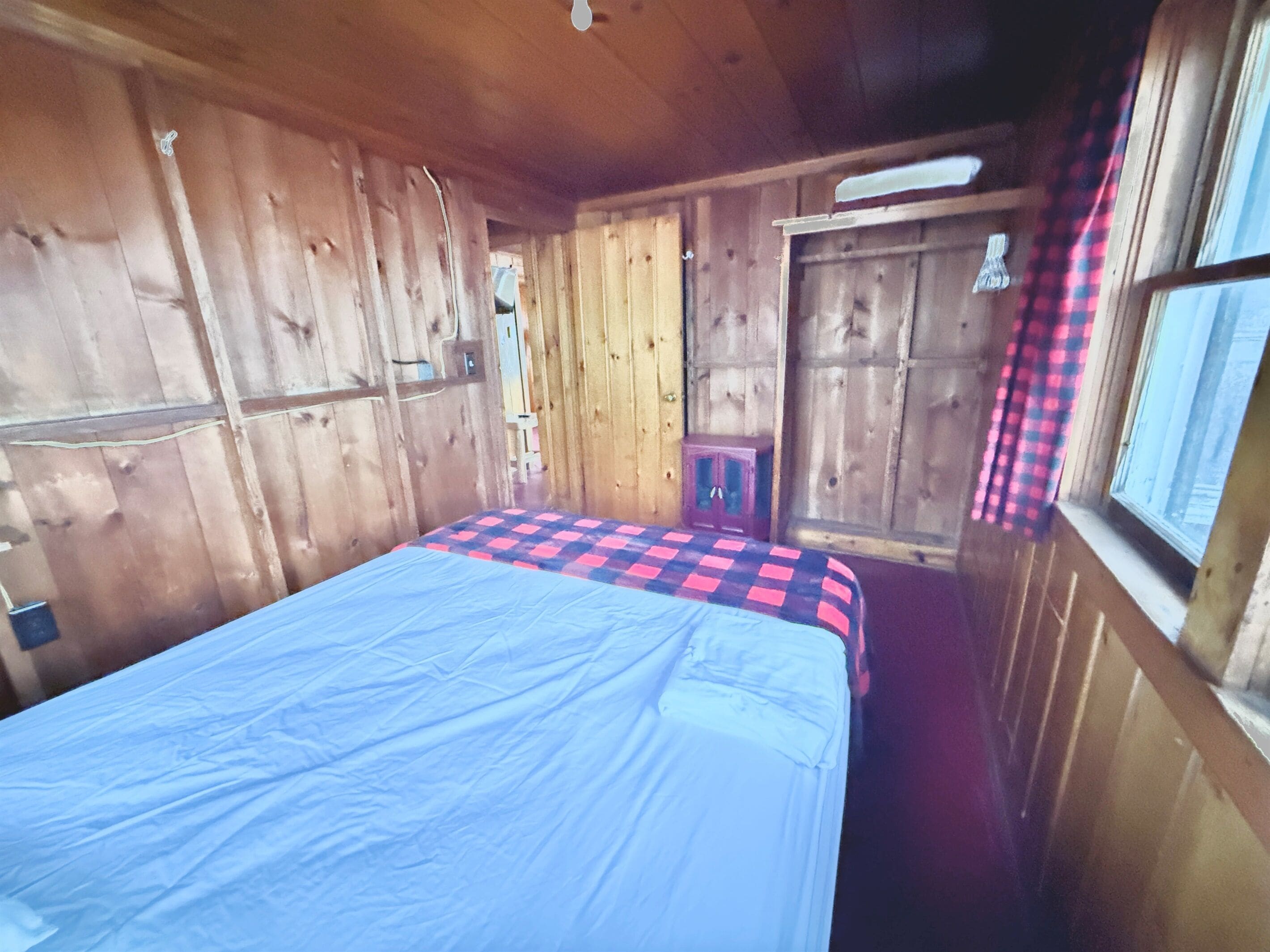 A wooden room with a bed, a window with a checkered curtain, and a door.